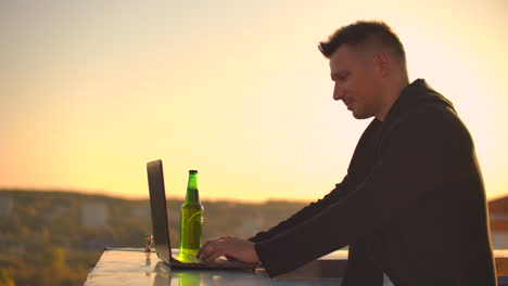 A-male-stockbroker-freelancer-stands-on-a-rooftop-at-sunset-with-a-laptop-and-types-on-a-keyboard-with-his-fingers-looking-at-the-cityscape-from-a-bird's-eye-view.